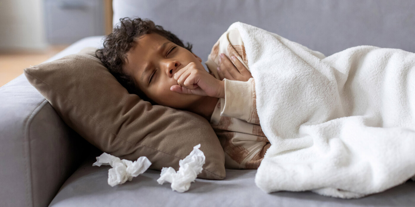 A young boy lies on a couch covered with  used tissues and coughs