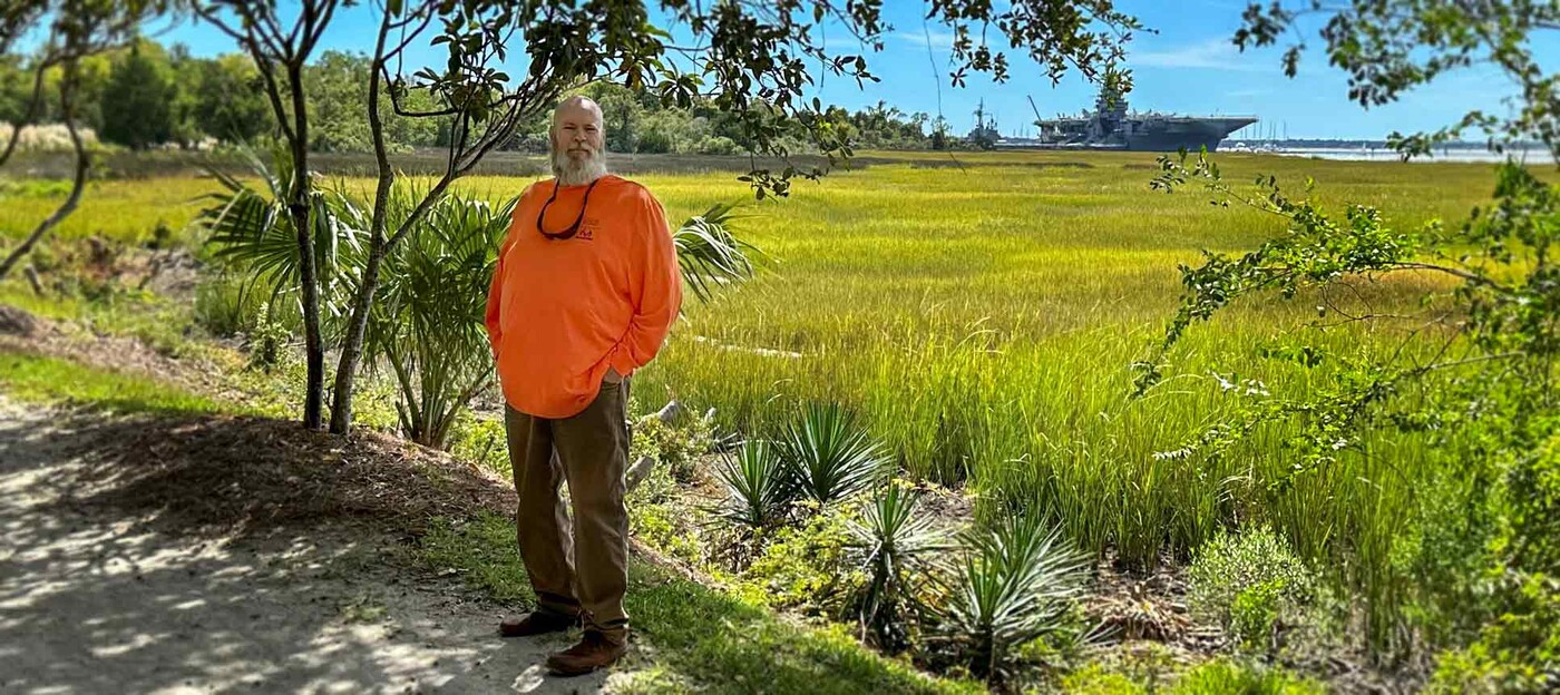 John White stands on a path with a marsh and a boat in the background