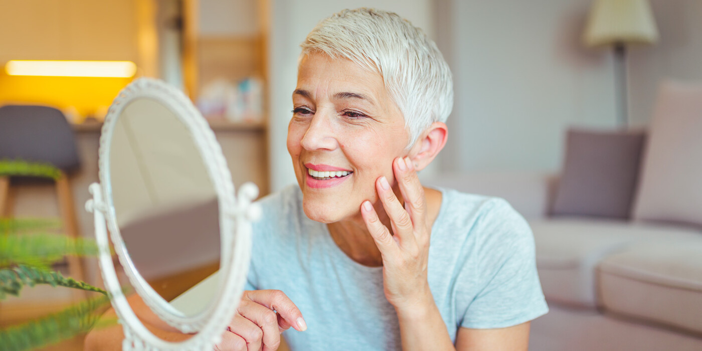 A woman smiles as she touches her face while looking in a mirror