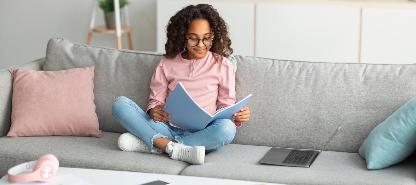 A girl reads on a couch next to an open laptop