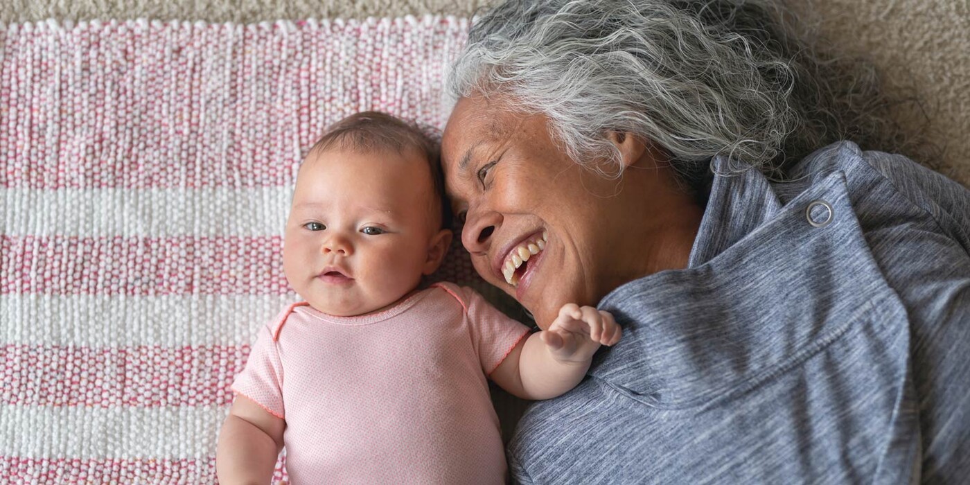 A grandma smiles at her grandchild as they cuddle on a blanket on the floor