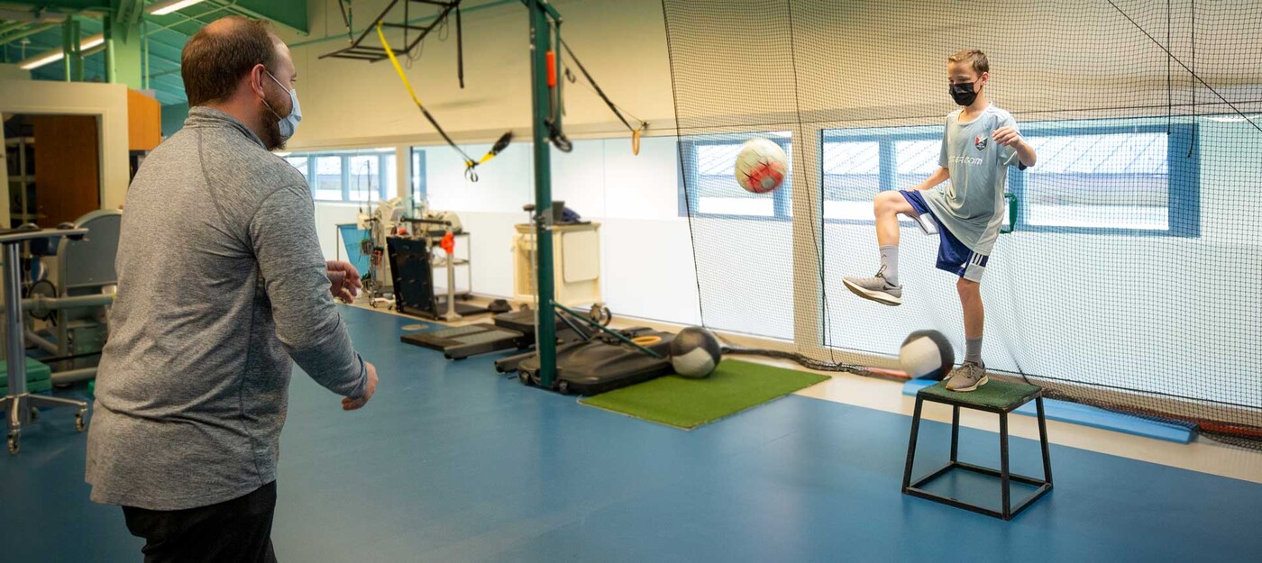 A physical therapist works with a young patient as he practices kicking a soccer ball while balanced on a raised platform.