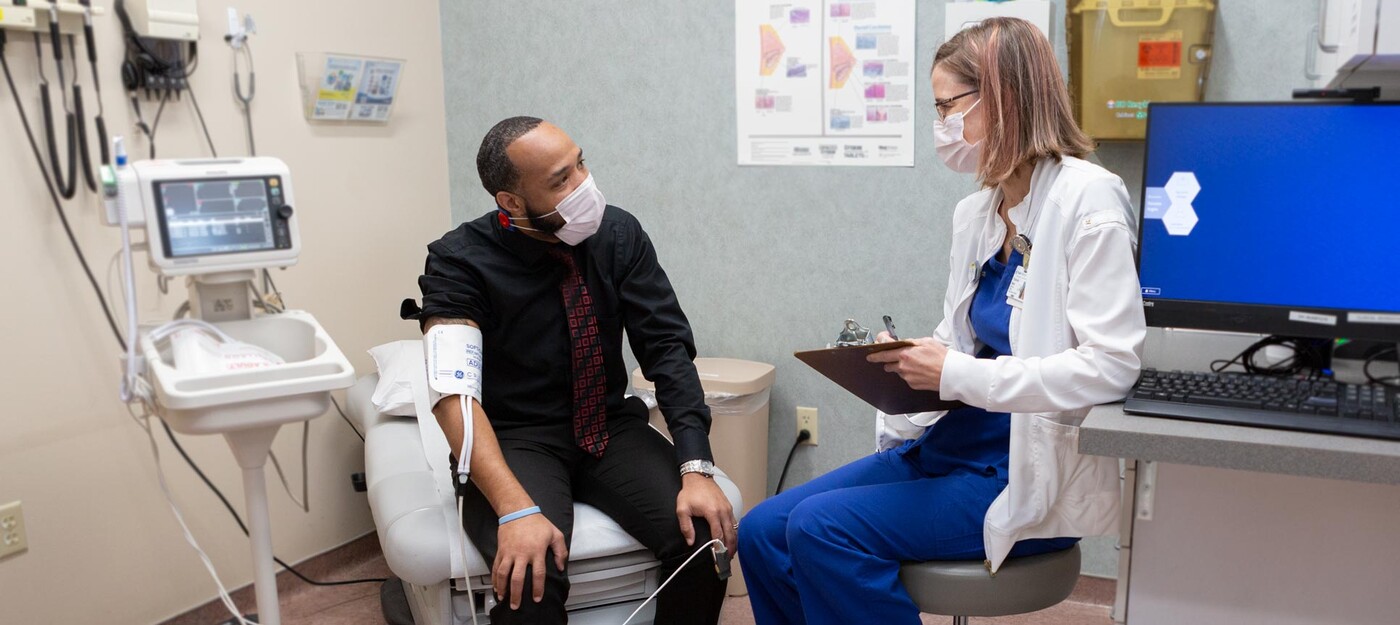 A patient sits with a blood pressure monitor on as he talks to a medical provider in a clinic room