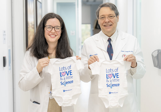 Two doctors hold up onesies that read "Lots of love and a little science"