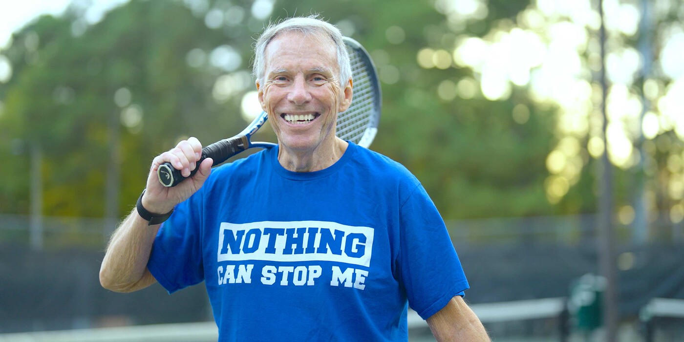 Joe Mesiano holds a tennis racket over his shoulder on a tennis court