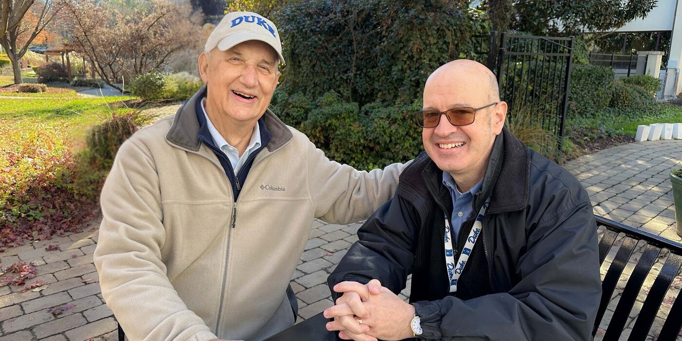 Two men sit outside at a table, smiling at the camera
