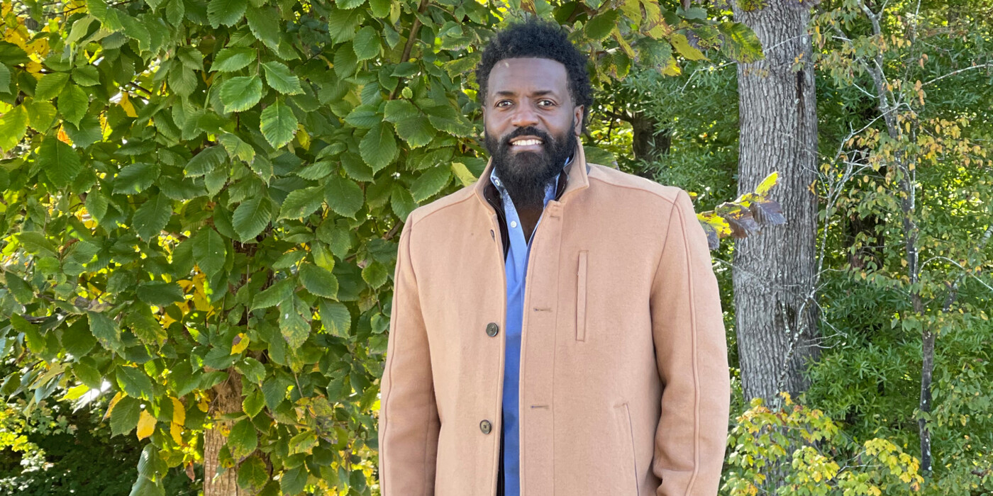 Stefan Rucker, wearing a tan coat, smiles in front of some trees.